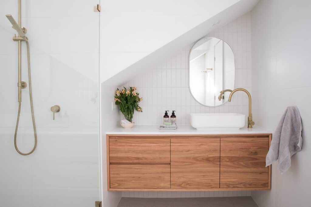 Salle de Bain Bois Laiton Moderne et Lumineuse Salle de bain moderne sous pente avec vanité flottante en bois clair, lavabo vasque blanc, robinetterie laiton et douche à l'italienne. Fleurs fraîches.