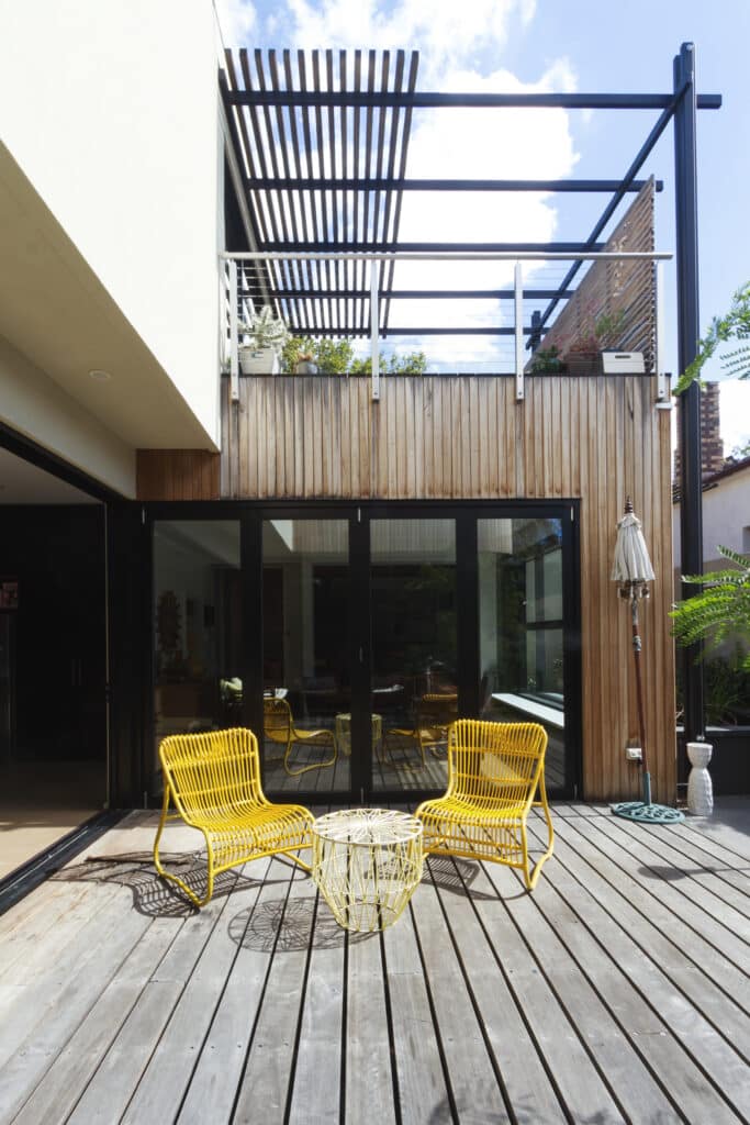 Terrasse moderne boisée et mobilier jaune design Terrasse en bois avec deux chaises jaunes et une table filaire, face à une façade moderne boisée et de grandes baies vitrées.